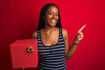 Young african american woman holding birthday gift standing over isolated red background very happy pointing with hand and finger to the side