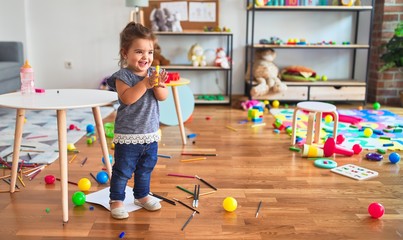 Beautiful toddler holding yellow pen standing around lots of toys smiling at kindergarten