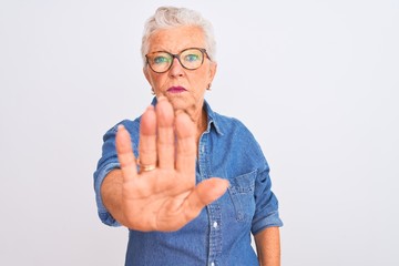 Senior grey-haired woman wearing denim shirt and glasses over isolated white background doing stop...