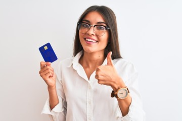 Young businesswoman wearing glasses holding credit card over isolated white background happy with big smile doing ok sign, thumb up with fingers, excellent sign