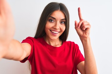 Beautiful woman wearing red t-shirt make selfie by camera over isolated white background surprised with an idea or question pointing finger with happy face, number one