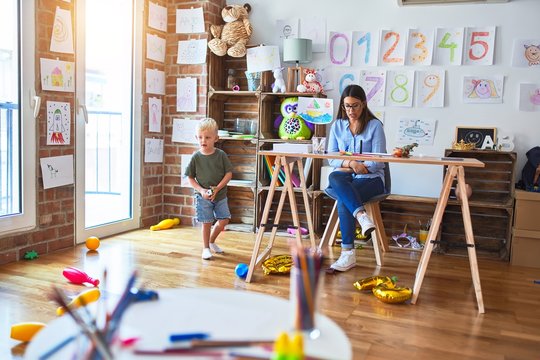Young caucasian child playing at playschool with teacher. Young woman sitting on the desk of the classroom