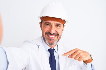 Middle age engineer man wearing helmet make selfie over isolated white background with surprise face pointing finger to himself