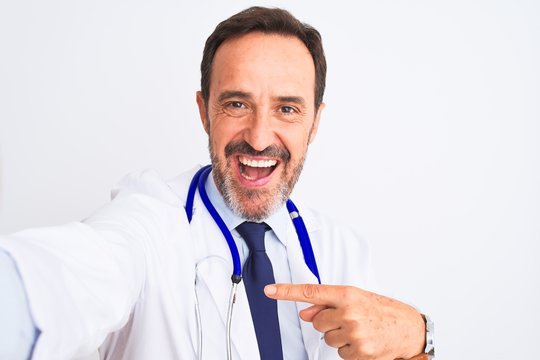 Middle age doctor man wearing stethoscope make selfie over isolated white background very happy pointing with hand and finger