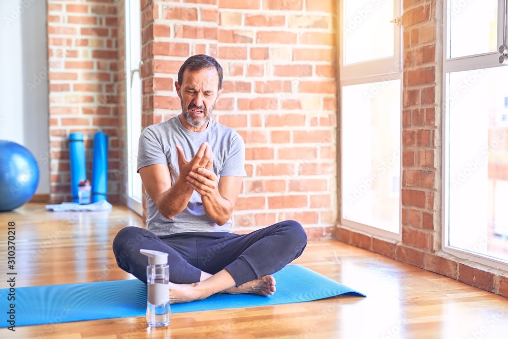 Poster Middle age handsome sportman sitting on mat doing stretching yoga exercise at gym Suffering pain on hands and fingers, arthritis inflammation