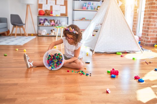 Beautiful toddler wearing glasses and unicorn diadem sitting on the floor playing with building blocks at kindergarten