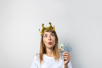 Young woman with a crown on her head and a surprised face holds stack of money on light background....