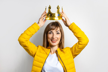 Young woman in a yellow jacket puts a golden crown on her head on a light background. Emotion...