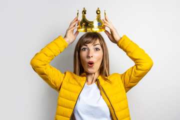 Young woman in a yellow jacket puts a golden crown on her head on a light background. Emotion...