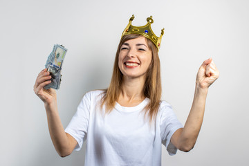 Young woman with a crown on her head holds a stack of money and celebrates very joyfully on a light background. Emotion laughter, surprise, kiss, shock. Queen, luck, wealth, gain, victory, bet. Banner