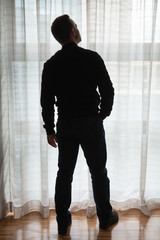 Young man stands near bright window with tulle and looks up