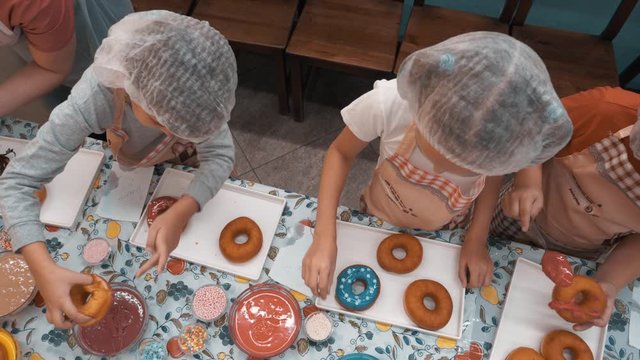Top view kids cooking donuts with colorful glaze on bakery master class. Overhead children preparing doughnut in culinary class in bakery shop. Culinary education for girls