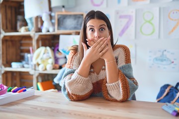 Young beautiful teacher woman wearing sweater and glasses sitting on desk at kindergarten shocked...