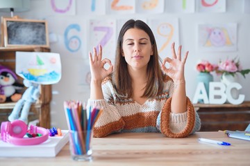 Young beautiful teacher woman wearing sweater and glasses sitting on desk at kindergarten relax and...