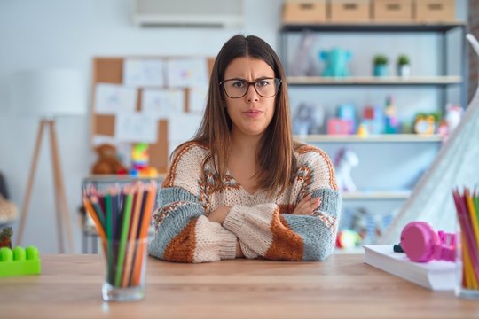 Young Beautiful Teacher Woman Wearing Sweater And Glasses Sitting On Desk At Kindergarten Skeptic And Nervous, Disapproving Expression On Face With Crossed Arms. Negative Person.