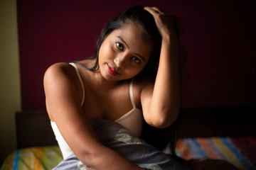 An young and attractive Indian Bengali brunette woman in white sleeping wear sitting on a bed inside in her room. Indian lifestyle.