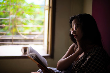 A young and attractive Indian Bengali brunette woman in white sleeping wear with a coffee/tea cup reading book on a bed in front of a window inside in her room. Indian lifestyle.