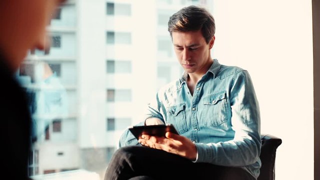 Attractive millennial young business man is using tablet in the office against the background of a window and glass buildings. Camera's coming from behind man's back. Tracking shot in slow motion.