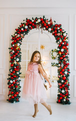 A little girl in a elegant pink dress is dancing in a room against the background of a Christmas arch.