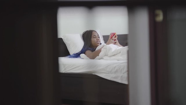 Happy smiling African American girl with dreadlocks using smartphone in bed. Teenager surfing Internet at night in her room. Social media addiction, lifestyle.