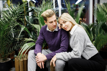 Fototapeta premium Cute blond guy and girl sitting among tropical plants in big store. Shop for decor