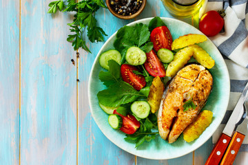 Dietary and healthy nutrition. Grilled salmon steak and baked potatoes with fresh vegetable salad on a wooden table. Top view on a flat background. Copy space.