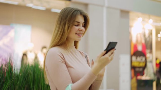 Zoom in panning shot of happy young woman standing with shopping bags in mall and browsing through pictures or text messages on cellphone