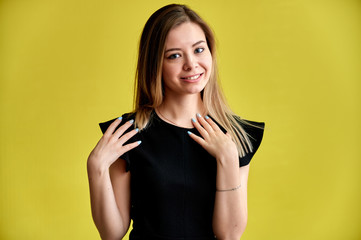 Close-up portrait of a pretty young smiling woman on a yellow background in a black dress with long straight hair. Standing right in front of the camera, Shows emotions, talks in different poses.