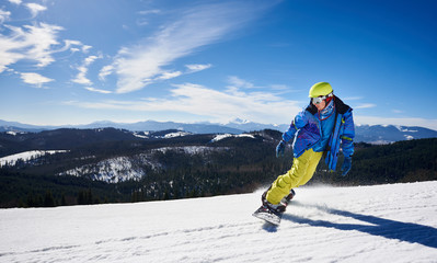 Fototapeta na wymiar Snowboarder man in goggles, helmet and warm bright clothing riding snowboard on background of blue sky and snowy mountains covered with spruce trees on sunny winter day. Sport and recreation concept.