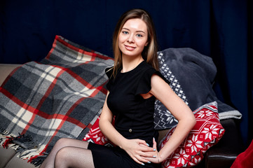Portrait of a beautiful girl with long hair in a black dress sitting at home on a sofa in a loft...