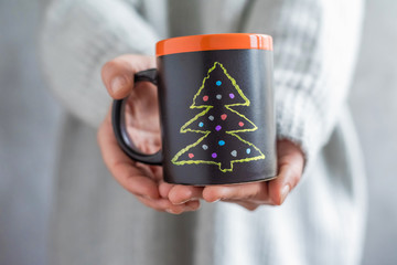 A girl in a huge knitted grey sweater holding a handmade slate black cup with painted Christmas tree. DIY
