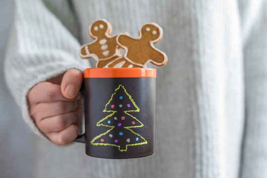 A girl in a huge knitted grey sweater holding a handmade slate black cup with painted Christmas tree and gingerbread men cookies. DIY. Concept