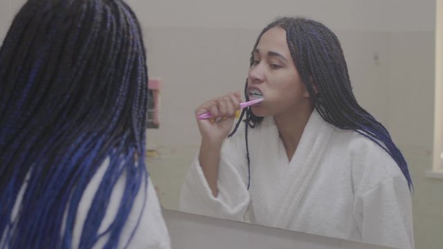Reflection in mirror of African American woman brushing teeth. Teenager getting ready in the morning in bathroom. Beauty, lifestyle, healthcare.