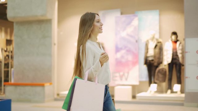 Beautiful young woman with shopping bags walking along mall, looking at window displays and smiling after successful shopping, three quarter length tracking shot