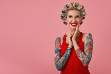 Cheerful young attractive blonde lady with tattooed hands smiling and looking dreamily upwards, foretasting upcoming party and preparing for it, standing against pink background