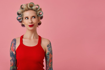 Indoor photo of attractive young blonde female with tattooed hands having curlers on her head and looking excitedly aside, wearing festive makeup while standing over pink background