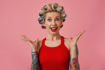 Close-up of amazed young attractive blonde woman with tattooes and evening makeup making hairdo and looking at camera with wide eyes opened, raising palms surprisedly over pink background