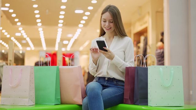 Tracking medium shot of young woman sitting on bench in mall surrounded by bags with purchases after successful shopping and browsing through photos or messages on cell phone