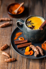 Golden milk in grey mug. Turmeric latte made with curcuma, cinnamon, anise. Healthy hot winter drink, natural, organic beverage. Close up, front view. Wooden rustic background. Raw roots as decor