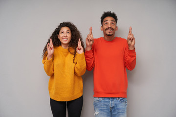 Indoor photo of positive young pretty dark skinned brunette couple crossing fingers for good luck and looking happily upwards, dressed in casual clothes over grey background