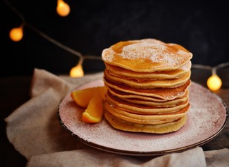 Pancakes with icing sugar and orange. Pancakes in a beautiful pink plate on a dark black wooden background. Delicious sweet food.