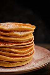 Pancakes with icing sugar and orange. Pancakes in a beautiful pink plate on a dark black wooden background. Delicious sweet food.