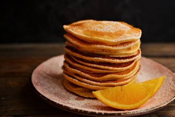 Pancakes with icing sugar and orange. Pancakes in a beautiful pink plate on a dark black wooden background. Delicious sweet food.