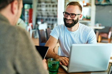 Fototapeta premium Attractive bearded man with glasses in a cafe working on laptop and chatting with a friend 