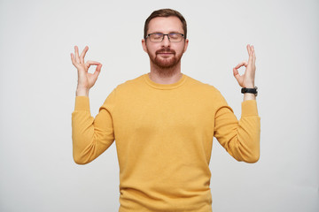 Pleasant looking young brunette bearded guy in eyewear raising hands with mudra gesture and keeping...