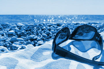 Close-up sunglasses on beach towel on pebbly sea beach. Trendy toning in color 2020 Classic Blue. Vacation concept.