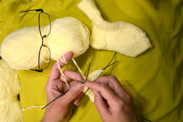Knitting needles with knitting thread in mom's hands close-up on a mustard color background. Two skeins of thread of light yellow color, knitted sock and glasses closeup.