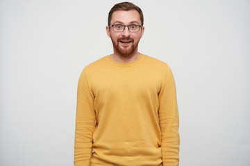 Suprised young brown haired pretty guy with beard wearing casual pullover and glasses while standing over white background with hands down, looking surprisedly at camera