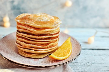 Pancakes with icing sugar and orange. Pancakes in a beautiful pink plate on a light wooden background. Delicious sweet food. Holiday pancakes and Christmas garland on the background.