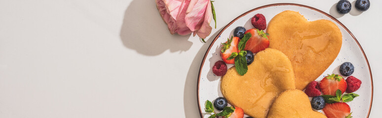 top view of delicious heart shaped pancakes with berries near rose on white background, panoramic...